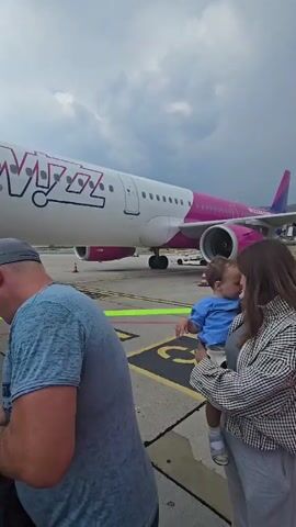 The video shows a group of people boarding an airplane. A jet bridge connects the terminal to the aircraft. A Wizz Air airplane is visible on the tarmac. People are walking up the stairs of the jet bridge, carrying luggage. The sky is cloudy.
