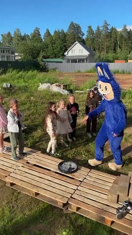 A group of children, some standing on a wooden platform, watch as a person in a blue cartoon character costume, possibly a hedgehog, uses a stick to poke at a black, circular object on the platform. The character is interacting with the children, pointing and gesturing. The scene appears to be outdoors in a grassy area with trees and houses in the background. The sky is clear and blue.