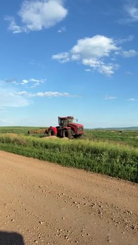 A bright red tractor is parked in a grassy field under a blue sky with white clouds. A dirt road is in the foreground. There are some mounds of dirt or soil behind the tractor.