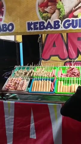 The video shows a food stall selling various types of skewered food items. The stall has a yellow banner with images of food and text that reads "KEBAB GURIH KEBAB MA". Below the banner, there are trays filled with rows of skewered items. The items appear to be sausages of different colors and sizes, as well as some round, breaded items. There are at least two individuals visible at the stall, one of whom is a young man wearing a black t-shirt, seemingly attending to the food. Another person, partially visible, is facing the stall. The stall itself is set up outdoors, likely at a night market or fair, indicated by the lighting and the red and white striped fabric hanging below the display. The overall impression is a busy food vendor preparing and displaying food for sale.