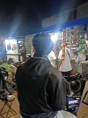 The video shows a nighttime scene at an outdoor market. A person wearing a black jacket is in the foreground, facing away from the camera. In the background, there are several stalls with goods displayed, illuminated by artificial lights. Various people are visible moving around the market. A row of parked motorcycles is also present. One stall appears to be selling packaged goods, such as snacks or cigarettes, hanging from displays. Another stall seems to have produce on display. The ground is dirt or gravel. The overall impression is of a bustling, dimly lit marketplace.