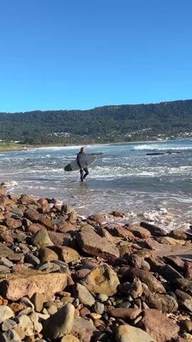 A surfer, dressed in a black wetsuit, is walking from a rocky shore into the ocean. The surfer is carrying a light-colored surfboard under their arm. The water is clear and turquoise, with small waves breaking. In the background, there's a mountain covered with trees and a few houses scattered along the coastline. The sky is clear and blue. The foreground shows a collection of rocks, varying in size and color, mostly reddish-brown, on the sandy beach.