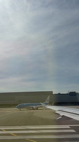 The video shows an airport scene viewed from inside a plane. The camera appears to be positioned by the window. We can see the wing of the plane the camera is inside, and another KLM airplane is stationary on the tarmac. A large building, likely part of the airport terminal or facilities, is visible in the background. The sky above is bright with light clouds. The ground is marked with yellow lines, and the overall setting is indicative of a commercial airport environment.