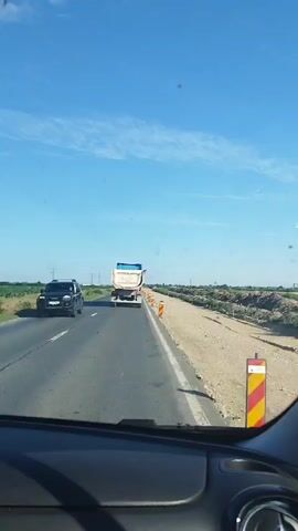 A white dump truck drives on a paved road under a clear blue sky. The truck is heading away from the camera. The road has white lane markings. To the right of the road, there is a dirt shoulder with orange and yellow striped traffic cones. In the distance, there are fields and utility poles. The video is shot from inside a vehicle, showing the dashboard in the foreground.