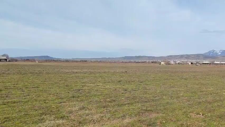 The video shows a wide shot of a grassy field with sparse vegetation under a cloudy sky. In the distance, a small village or cluster of houses is visible, nestled at the foot of a large mountain range capped with snow. Three people are walking through the field.