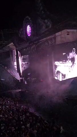 The video captures a vibrant outdoor concert at night. A large, intricately designed stage dominates the scene, featuring a prominent circular screen with purple lighting. The stage is adorned with numerous lights and pyrotechnic effects, including bursts of fire and green lasers that cut through the smoky atmosphere. Below the stage, a massive crowd of people, mostly visible from the chest up, is gathered. They are facing the stage, with many holding up their hands and phones, suggesting they are actively engaged with the performance. The overall impression is one of a high-energy event with a dramatic visual display and an enthusiastic audience.