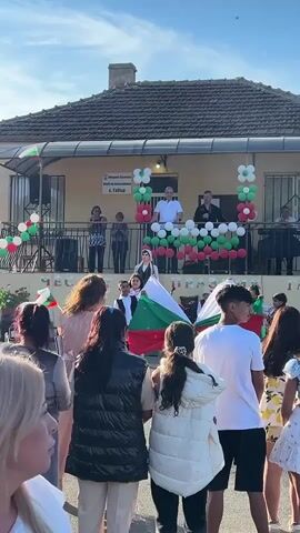 A group of people are gathered outdoors in front of a building with a covered porch, which is decorated with balloons in red, white, and green. Several children are on the porch, holding Bulgarian flags. A woman in the foreground is looking to her left, her face partially visible. A young child in a white outfit is walking away from the camera. The sky is blue with some clouds. The overall scene appears to be a public event or celebration.