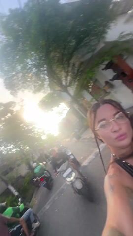 A first-person-perspective video shows a woman on a scooter, holding a bottle of Coca-Cola. She is fair-skinned, with long, braided brown hair and gold wire-rimmed glasses. She is wearing a black, sequined top with a plunging neckline and black gloves. She smiles at the camera. The person holding the camera has a large tattoo on their arm. They are sitting on a scooter with a black helmet and green accents visible. The video appears to be taken in an outdoor, urban area, with houses, trees, and other parked scooters visible. In the background, a large sign advertises "The Best Deep Tissue Massage." The video has bright lighting, with some lens flare from the sun.