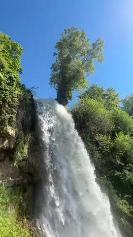 The video shows a scenic view of a waterfall. Water cascades down from a rocky cliff, creating a large spray at the bottom. Lush green vegetation surrounds the waterfall, contrasting with the clear blue sky visible above. The camera slowly pans down to capture the full length of the waterfall and the dense greenery at its base.