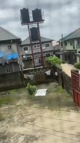 The video shows a courtyard in what appears to be a residential area. The courtyard is surrounded by concrete walls and buildings. In the courtyard is a small tree and a piece of white material on the ground. Laundry is hanging on a line strung along the courtyard walls. Outside the courtyard is a street with more buildings. In the distance, there is a tall metal structure holding water tanks. The sky is overcast.