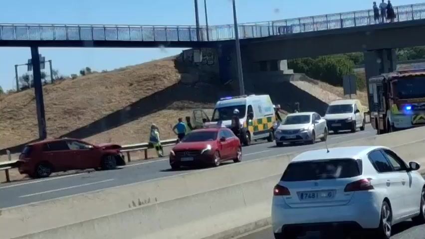 The video shows a multi-vehicle accident on a highway. Several emergency vehicles are present, including ambulances and fire trucks with flashing lights. Law enforcement officers and paramedics are attending to the scene. There are multiple damaged cars and vans, suggesting a significant collision. People are seen standing near the vehicles, some appearing to be providing assistance or observing the situation. An overpass is visible in the background, with a few individuals standing on it. The overall environment appears to be a sunny day on a busy roadway.