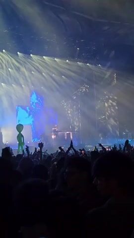 A concert crowd is visible in the foreground with their hands raised. Stage lighting illuminates a DJ booth with a person operating equipment. A large screen behind the DJ displays abstract blue graphics and a green alien figure. The environment appears to be an indoor arena filled with haze and bright spotlights.