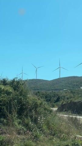 The video shows a scenic view from a moving vehicle. The focus is on a hill dotted with wind turbines against a clear blue sky. The road curves in the foreground, with vegetation and a sign visible along the side. The camera then pans upwards, capturing the landscape and the view through the car window.