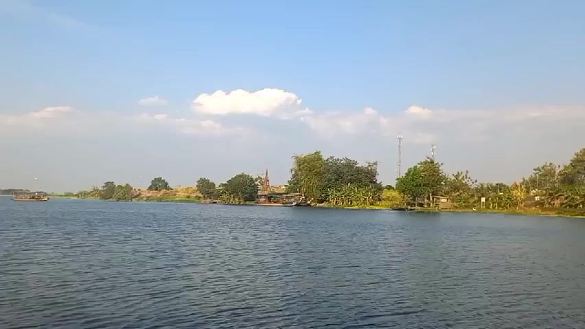The video is shot from the perspective of someone on a boat traveling down a wide river. The river is calm and reflects the blue sky, which has a few scattered white clouds. On the left bank of the river, there are some trees and what appear to be houses or buildings. A few boats are visible in the distance on the river. On the right bank, there are more trees, some of which are very tall and leafy. There are also some smaller structures along the bank, possibly houses or sheds. There are two cell towers visible on the right bank, one taller than the other. In the foreground on the left side of the screen, a person wearing a dark blue shirt and a black hat is visible, looking out at the river. In the foreground on the right side of the screen, a person wearing a dark shirt is partially visible. The overall impression is a peaceful, rural scene.