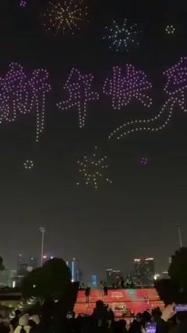 At night, a drone show illuminates the dark sky with colorful patterns and Chinese characters. The display includes purple, blue, and multicolored “firework” bursts. The characters are lit in purple and white, while smaller bursts of light appear around the central display. Below, a crowd of people stand on illuminated red stairs, likely watching and filming the show. The background features a cityscape with buildings and streetlights. The atmosphere is celebratory, with sounds of the crowd audible.