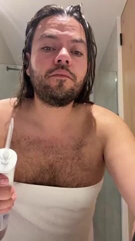 A man with wet brown hair, a beard, and a hairy chest covered by a white towel is in a bathroom. He is holding a white water flosser and using it on his teeth. He is looking up. A shower is visible in the background.
