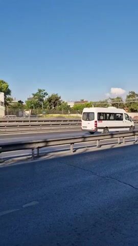 The video shows a view from a moving vehicle looking out at a highway. The sky is clear and blue. On the highway, there are several vehicles including cars and vans. Some buildings and trees are visible in the background. There is a concrete wall with large billboards along the right side of the road. A metal guardrail separates the highway lanes. Traffic signs with text are also visible. The road surface is asphalt. The video appears to be taken during daytime.