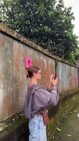 A woman wearing a grey sweatshirt, blue jeans, and sunglasses is taking a photo with her pink phone. She has her hair pulled back in a bun and a small brown purse attached to her hip. The woman is standing beside a mossy wall with a small pink art piece attached to it. She takes a picture of the art and then a picture of a large tree with light grey roots that look veiny. There is a lush green tree behind the wall in the background.