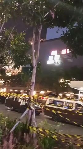 The video shows an outdoor scene at night. In the foreground, there are plants and a tree. Yellow and black caution tape is strung across the scene. In the background, there is a building with illuminated signs. There are also vehicles, possibly vans, with caution tape wrapped around them. Further back, there is a walkway or structure with railings and people can be seen moving around. Some areas appear to be roped off with red and white barriers. The overall impression is of an area that may be under construction or undergoing some form of restricted access or event.