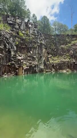 A drone shot pans across a quarry lake. The water is a bright, unnatural green. Sheer, dark rock walls rise dramatically from the water's edge, with jagged formations and vertical striations. Trees and brush grow along the top of the quarry rim, and the sky above is blue with scattered white clouds and bright sunlight.