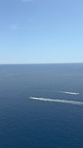 The video shows a daytime view of a deep blue ocean under a clear, light blue sky. Two motorboats are speeding across the water, each leaving a distinct white wake behind them. A bird is flying in the sky.