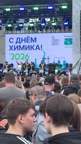 A crowd stands watching a marching band of approximately six drummers on a large outdoor stage. The drummers are dressed in marching band uniforms. Behind the band, a large LED screen displays "С ДНЁМ ХИМИКА!" in large green letters, which translates to "Happy Chemist's Day!". Below, the number "2026" is displayed. The event seems to be sponsored by PhosAgro, as their logo appears on the right side of the screen with the tagline "НА СЕВЕРЕ - ЖИТЬ!". Stage lighting is visible above the screen, and many people in the crowd are holding up their phones to record the performance.