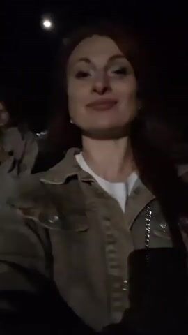 This is a short video taken at night. In the foreground, a woman with long red hair, wearing a tan jacket over a white shirt, is holding a black object, likely a phone or camera, and looking into it. She is smiling and making a peace sign with her right hand. Behind her and to her left, a person in a blue and white plaid shirt is partially visible. Further back and to the right, another person in a tan shirt and light-colored pants is also visible. The environment appears to be outdoors, possibly a street or park, with some blurry lights in the background. The video is shaky, suggesting it was filmed while walking.
