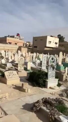 The video shows a large cemetery with numerous headstones of varying styles and colors, mostly white and beige. The grave markers are set in a sandy, dirt-covered ground. Some graves are adorned with dried plants. The cemetery is surrounded by a low wall and adjacent to buildings that appear to be residential or commercial. The sky is overcast, with the sun diffused.  No people are visible in the scene.