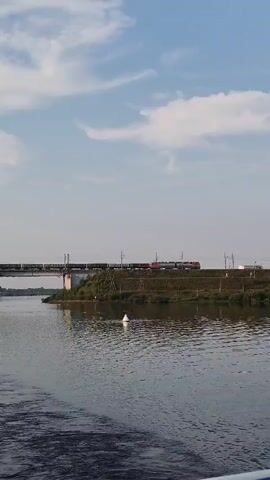 A train crosses a bridge over a body of water. The sky is partly cloudy.