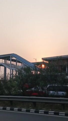 The video shows a road at sunset. A footbridge spans the road in the first shot. A white car drives past. Subsequent shots show more traffic on the highway, including a black SUV and another white car. The sky is pale with the setting sun. The road is marked with white lines and a guardrail. Greenery is present on the roadside. A curve warning sign is visible.
