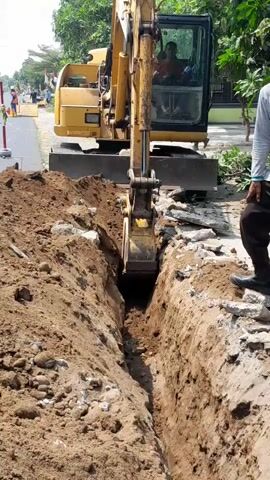 An excavator is digging a trench along a road. The excavator is yellow and has a person in the cab. A man in a light-colored shirt and dark pants is standing on the right side of the trench, pointing towards the excavator. The trench is filled with dirt and rocks. There are trees and houses in the background.