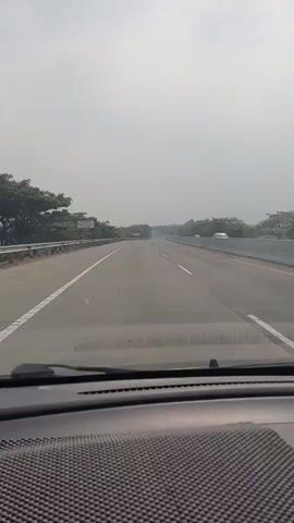 The video shows a first-person perspective from inside a car driving on a highway. The road ahead is clear with multiple lanes and is bordered by trees and a guardrail. The sky is overcast, creating a hazy, bright atmosphere. There are no other vehicles visible in the immediate foreground, though some may be present further down the road. The dashboard of the car is visible at the bottom of the frame.
