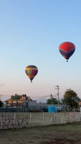 The scene shows two hot air balloons floating in the sky. The balloon on the left is multicolored with horizontal stripes, while the balloon on the right is primarily red with blue and gray sections. The balloons are seen against a backdrop of a pale blue sky transitioning to a slightly pinkish hue near the horizon. Below the balloons, there is a view of a fenced-off area with patchy grass. A building with signage that includes the word "HOTEL" is visible in the background, along with trees, power lines, and other buildings. There appears to be some activity near the fence, with a few figures and a white vehicle visible, but they are relatively small in the frame.