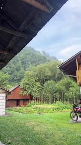 The video shows a rural backyard scene during a light rain. Two wooden structures, likely houses or sheds, are visible on the left and right sides of the frame, with a sloped roof in the foreground partially obscuring the sky. A red motorcycle is parked on the right side of the yard near one of the wooden structures. A green lawn and a garden area are in the mid-ground, leading up to a dense forest covering a hillside in the background. The sky is cloudy, with visible streaks of rain falling. The overall atmosphere is serene and natural.