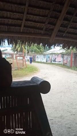The video begins with a view from under a thatched roof, looking out onto a dirt road. A person in a blue shirt is walking away from the camera on the right side of the road. To the left of the road, there is a playground with some equipment. Further down the road, there are buildings and a wall with graffiti. A young boy in a white shirt and black pants walks into the frame from the right and walks past the camera. The scene appears to be outdoors in a rural or semi-rural setting.