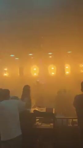 The video shows a scene in what appears to be a nightclub or concert venue. The space is dark, with blue and white laser lights flashing and moving across the room. The beams of light cut through fog or haze, creating visible shafts in the air. There are people in the room, mostly silhouetted, suggesting they are standing or dancing.  The focus is on the light show, and the environment suggests a lively, energetic atmosphere.