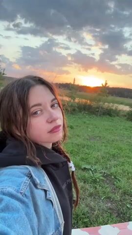 A woman with brown hair, styled in a braid, smiles as she takes a selfie outdoors at sunset. She is wearing a denim jacket over a black hoodie with the words "Great Mo" visible on it. The background features a grassy field with some trees and a body of water, all bathed in the warm glow of the setting sun against a cloudy sky.