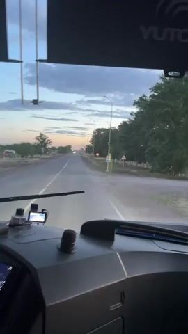 The video is a first-person view from inside a vehicle, looking out through the windshield at a road. The sky is overcast with clouds and hints of sunset. The road is paved and appears to be a highway with streetlights and trees lining the sides. There are other vehicles visible in the distance. The dashboard of the vehicle is visible in the foreground, with various buttons, screens, and a small camera mounted on the windshield. The scene suggests the vehicle is in motion, traveling along the road.