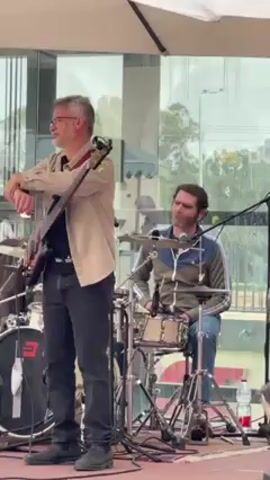 The video shows two men playing instruments in an outdoor setting. The man on the left is playing a bass guitar and is wearing a light brown shirt, black pants, and dark boots. He has gray hair and glasses. The man on the right is playing drums and is wearing a green and blue jacket, blue jeans, and white sneakers. He has dark hair and a beard. They are positioned in front of a building with large windows. The weather appears to be overcast. There is a microphone in front of the drummer. The background is slightly blurred.