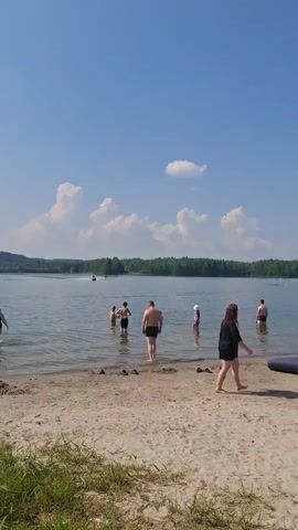 A bright, sunny day at a beach with a clear blue sky and scattered white clouds. A sandy beach slopes gently into a calm lake or bay. Several people are in the shallow water, some appear to be wading, while others are closer to the shore. In the middle ground, a jet ski is moving across the water. The background shows a dense line of green trees. On the right side of the beach, a dark blue inflatable object is visible. The overall scene depicts a relaxed recreational activity on a warm day.