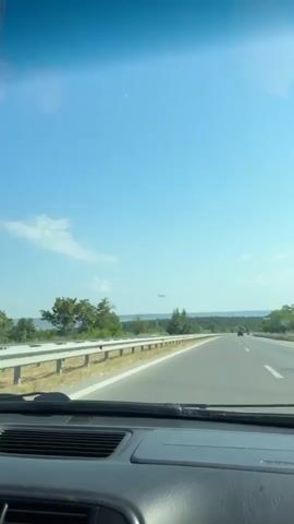 A first-person view from inside a car shows a highway with a clear blue sky overhead. A metallic guardrail runs along the side of the road, separating it from a grassy area with trees. In the distance, a few other cars are visible on the highway. The road curves gently to the right. The dashboard of the car is visible in the foreground.