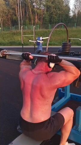 The video shows a man performing bicep curls with a barbell on an outdoor gym machine. He is shirtless, wearing black gloves and white headphones. He is seated and has his back to the camera. The man's skin appears reddish, possibly from sunburn or exertion. The gym equipment is blue and metal. In the background, there are trees and a fence. The lighting suggests it is daytime, possibly late afternoon or early evening.