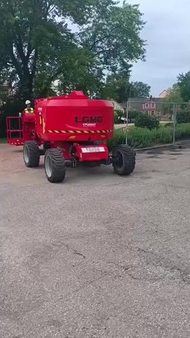 A person wearing a hard hat and a yellow vest stands on the platform of a large, red cement mixer with large, rugged tires. The cement mixer has "L.CMG STOGRENT" written on its side. The machine is on a paved surface, possibly a road or parking lot, with trees and buildings visible in the background. The video shows the cement mixer moving slightly to the left and right.