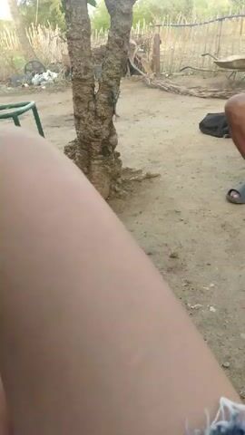 A video shows a person's leg in the foreground, with the camera positioned on the ground. Behind the leg, a tree with a rough, textured trunk is visible. In the background, there is a dirt area with some debris, a green chair, and another tree with leaves and branches against a blue sky with clouds. A palm tree is also visible.