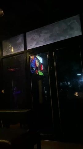 A dark interior space, possibly a club or bar, features a large window or doorway. Within the window frame, a television screen displays a brightly colored music video with a person holding what appears to be a weapon or instrument. The music video's visuals include bold patterns and color gradients. The overall environment seems dimly lit and noisy.