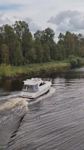 A white motorboat travels on a dark body of water, leaving a wake behind it. Trees and vegetation line the shore of the waterway. The sky is cloudy. The video shows the boat from an elevated, rear-facing perspective.