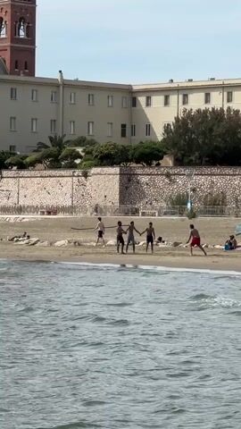 The video shows five young men playing soccer on a beach. The men are dressed in swimming trunks or shorts and are spread out across the sand near the water's edge. There are some other people sunbathing or relaxing on the beach. Behind the beach is a stone wall, and beyond that, a large, light-colored building with a tower is visible. The water is choppy, and the sky is bright.
