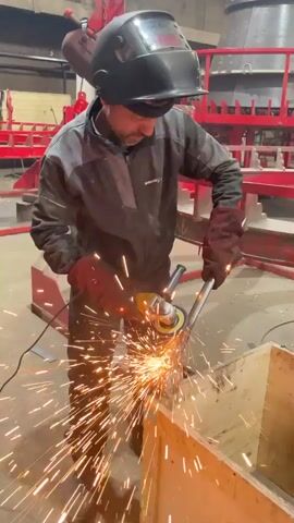 A man is standing in a workshop, wearing a dark gray jumpsuit, a black welding helmet, and maroon gloves. He is using a yellow angle grinder to smooth a round, metal object held in a wooden crate. Sparks fly as he grinds. In the background, red metal frameworks can be seen, suggesting an industrial setting. A cord connected to the grinder is visible near his feet.