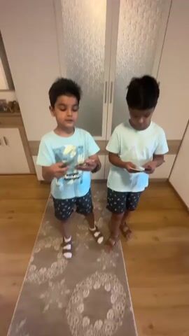 Two young boys are standing in a hallway and counting cards. The boy on the left is wearing a light blue t-shirt with a graphic print and dark patterned shorts. He is holding several cards in his hands and is counting them. The boy on the right is wearing a solid light blue t-shirt and dark patterned shorts. He is holding a few cards in his hand and appears to be watching the other boy. They are both wearing sandals. The hallway has wooden floors and a rug with a circular pattern. In the background, there are two large white closet doors with textured glass panels.