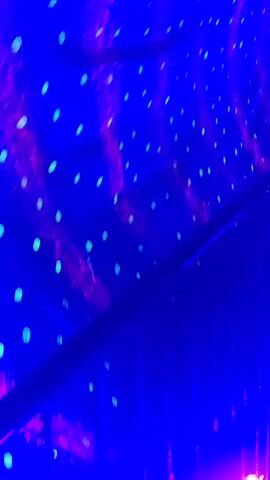 The video captures a person walking through a neon-lit, blue-toned tunnel that appears to be an attraction or a funhouse. The tunnel is decorated with numerous small, glowing dots of light, primarily teal and pink, creating a psychedelic and immersive atmosphere. Some areas show a person's silhouette, blurred by motion, suggesting movement through the space. The lighting is dramatic and enhances the sense of depth and disorientation. The person is wearing light-colored clothing, which stands out against the dark and vibrant surroundings. There are also glimpses of architectural elements like staircases and railings, reinforcing the idea of a constructed environment designed for entertainment. The overall impression is one of playful exploration in a visually stimulating, almost otherworldly setting.